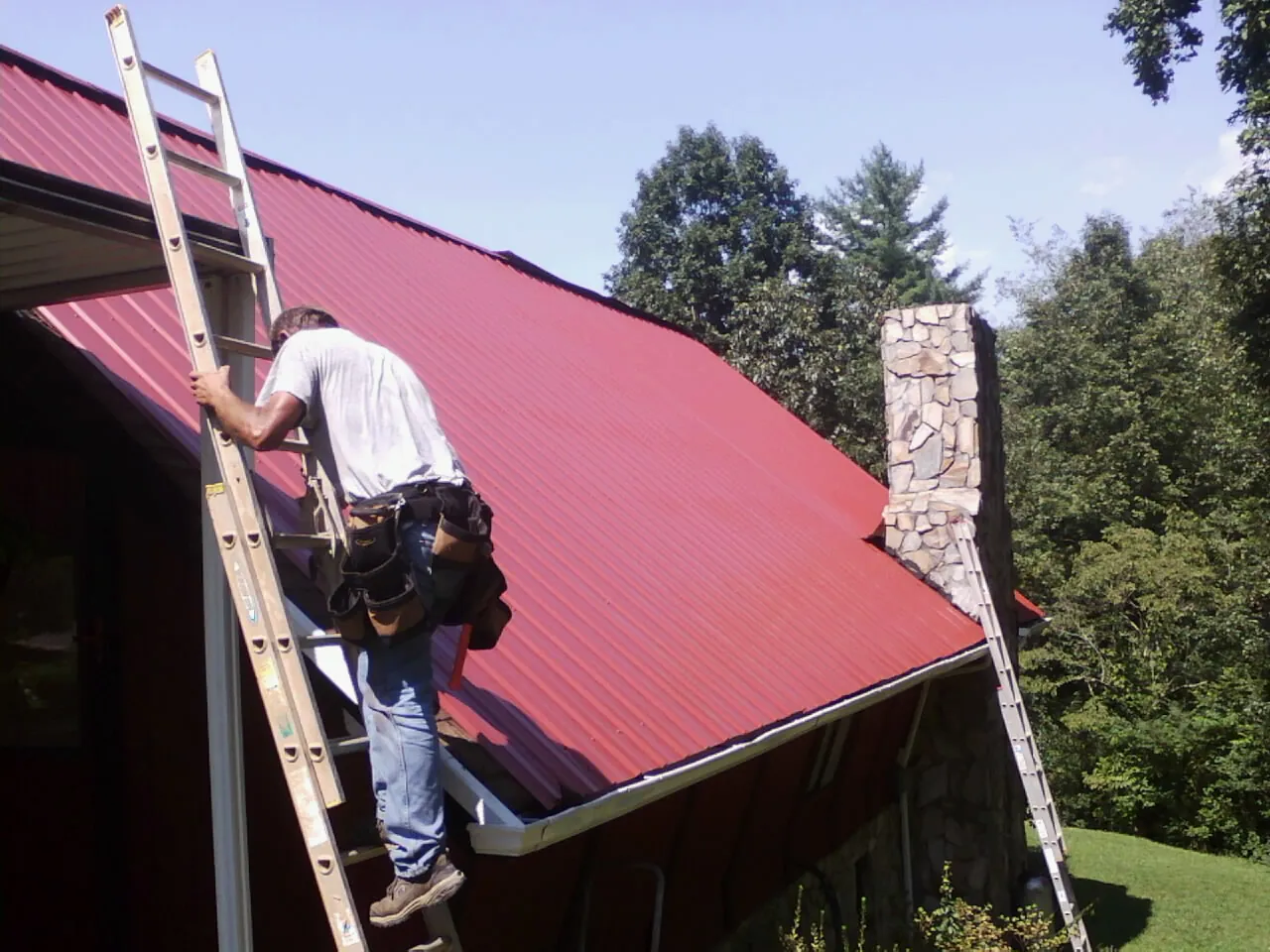 Roofer on a ladder beside a red metal roof and stone chimney — Soffit & Fascia Repair by Roofing Brothers in Wisdom